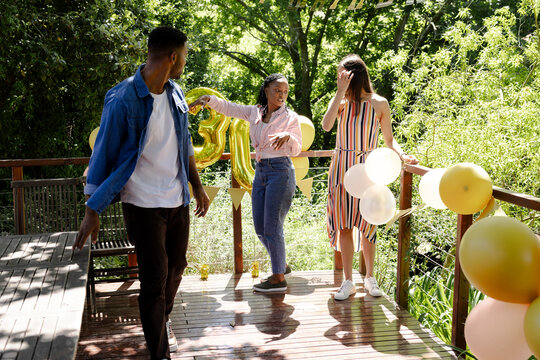 Diverse friends celebrating outdoors in garden with balloons, enjoying sunny day on wooden deck - Powered by Adobe