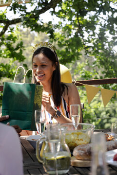 Smiling woman opening gift at outdoor birthday party with diverse friends and drinks