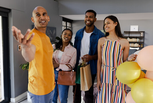 Holding gifts and balloons, diverse friends gathering at home, celebrating together happily