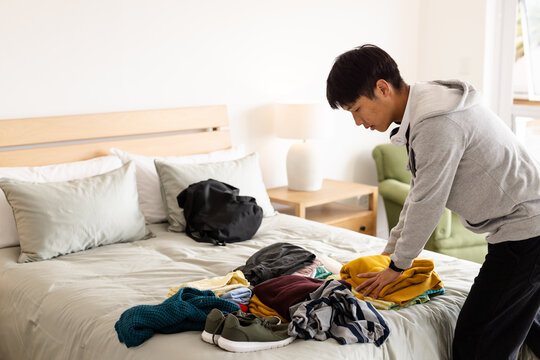 Man packing clothes on bed, preparing for travel with backpack nearby, at home