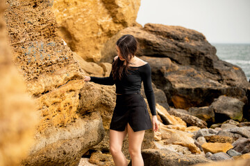 A young girl with long dark hair poses near a rock with the sea in the background. She is wearing a...