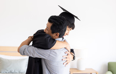 Proud father hugging graduate son in cap and gown at home, copy space