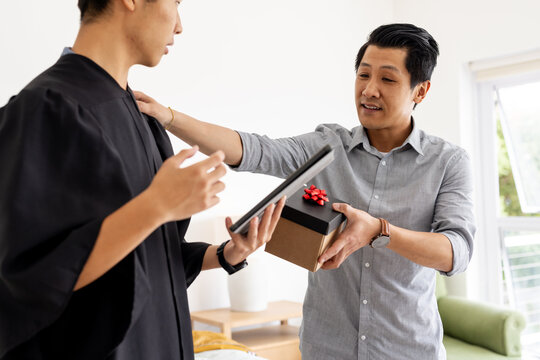 Proud father giving gift to graduate son holding diploma at home