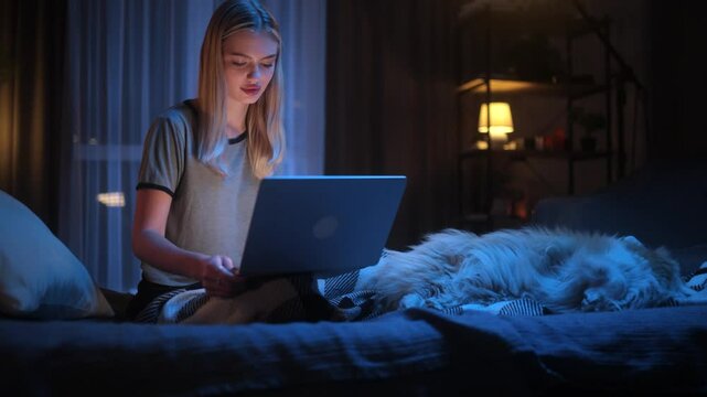 Young woman working on laptop in the evening while sitting on bed with her sleeping dog. Cozy and calm night atmosphere