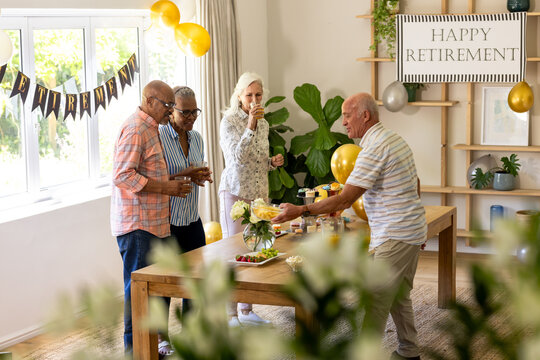 Celebrating retirement, senior diverse friends having drinks and snacks in decorated room, at home