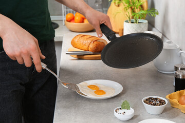 Man with frying pan putting tasty fried eggs onto plate in kitchen, closeup