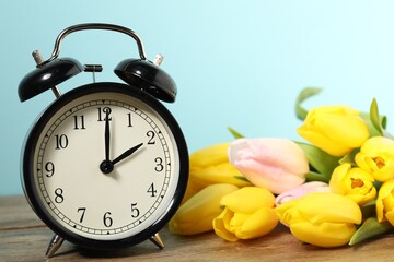 Spring time. Alarm clock and beautiful tulips on wooden table against light blue background