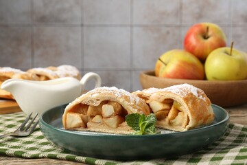 Pieces of tasty apple strudel with powdered sugar and mint on wooden table against grey tiled wall, closeup