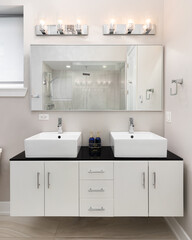 A bathroom detail with a floating white cabinet, vessel sinks sitting on a black marble countertop, and a view of the shower through the mirror.