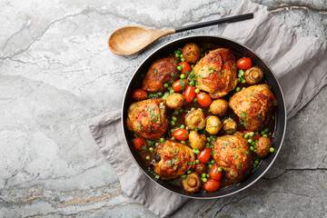 Fried chicken thighs with mushrooms, green peas, cherry tomatoes and herbs close-up in a frying pan on the table. Horizontal top view from above