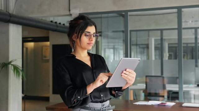 Latin Hispanic young professional business woman working on touchscreen computer. Middle eastern Indian businesswoman CEO holding digital tablet using tab application standing at workplace in office