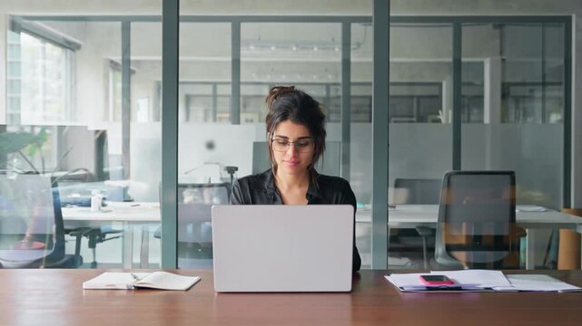 Young professional it specialist latin hispanic business lady working on laptop pc sitting at desk in modern office space. 30s middle eastern indian woman using computer technology app for work online