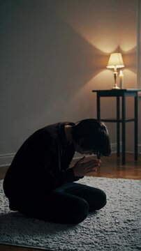 A dimly lit room with a person kneeling on a rug, hands covering face. Side angle captures a somber, introspective video scene with soft lighting.