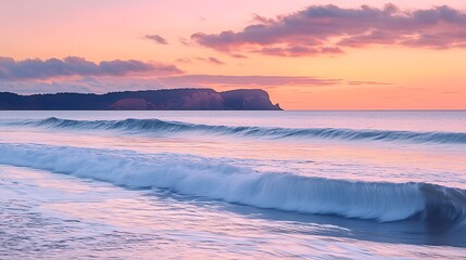Obraz premium Serene ocean waves rolling onto a beach at sunset with a cliff in the background.