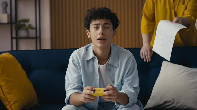 Teenager playing a video game, ignoring his mom showing his poor test result