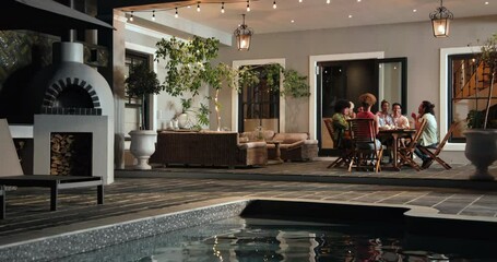 Tracking shot past swimming pool to happy diverse friends talking on terrace at dinner party