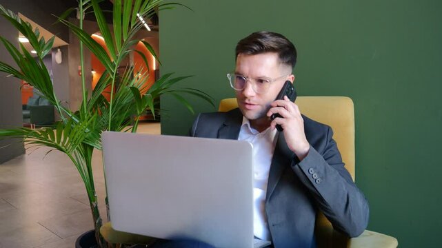Man in business attire uses smartphone while attending to laptop in a stylish office environment.