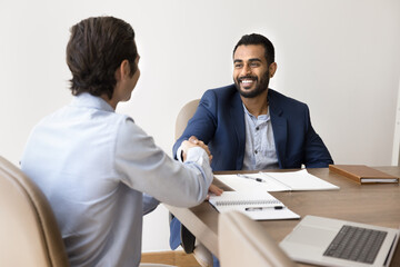 Two multiethnic professionals closing deal, exchanging firm handshake seated at conference desk,...