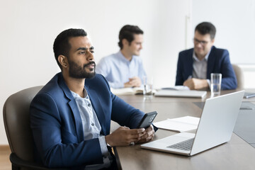 Indian businessman in suit sits at desk with laptop, hold smartphone looks into distance with pensive, thoughtful expression, ponder on answer, analyze received sms, wait for important business call
