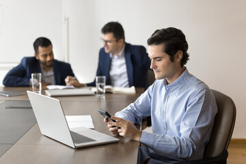 Focused businessman using smartphone sit at desk with laptop, check information, respond to messages, verifying data, reviewing updates or documents. Office worker workflow using modern wireless tech
