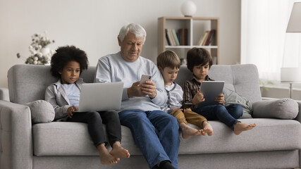 Quiet moment with digital devices. Intergenerational family senior grandpa multiracial early school age grandkids sit on cozy couch use different gadgets to watch education video text messages in chat