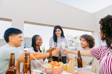 Group of Latin friends gathered for dinner, toasting with glasses on a terrace outside at sunset, celebrating