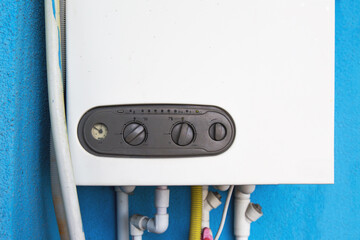 Gas boiler mounted on a blue wall with control panel and various pipes connected at the bottom. The boiler features adjustment knobs and a pressure gauge for regulating water heating
