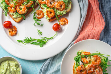 Healthy shrimp with avocado and arugula on toast for breakfast on a white plate on a light blue background. View from above. Flat layout.