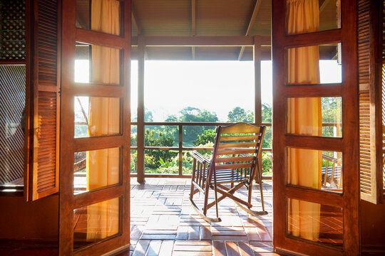 wooden glass doors opening out onto a balcony with a beautiful wooden rocking chair looking out over a tropical garden with bright sun shining onto the balcony, Arenal, Costa Rica, copy space
