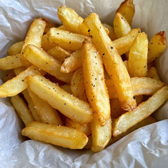 A serving of golden, crispy French fries arranged in a white paper container. The fries are evenly cooked with a light, golden-brown texture, resting on a marble surface that adds a clean