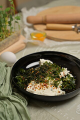 Parsley and dill greens with cottage cheese in a black bowl. Rolling pin, cutter, eggs, green towel and microgreens sprouts on kitchen table. Home cooking baking puffs