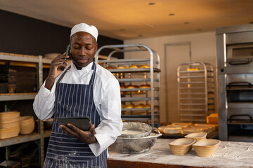 Baker in uniform using tablet and phone in busy bakery kitchen, copy space