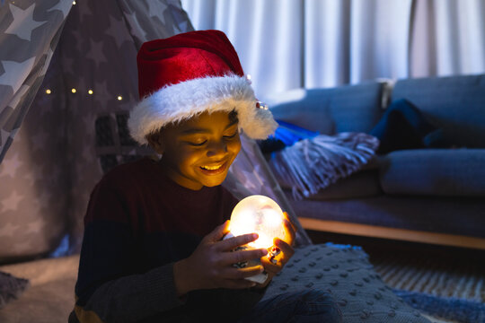 African American boy wearing Santa hat holding glowing orb, smiling joyfully at home