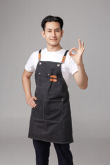Confident Asian man wearing a denim apron and white shirt, smiling and exhibiting a show Ok sign on...