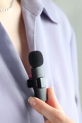 Woman putting lavalier microphone onto her shirt on light grey background, closeup