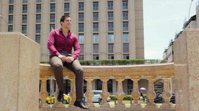 Young Executive sitting on a bench in front of a modern glass building, smiles looking at the camera in a positive and winning attitude.