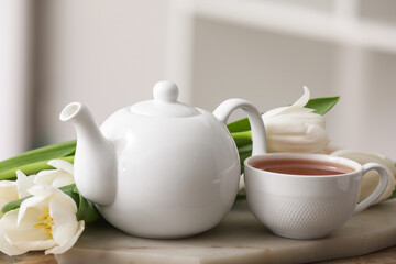 Board with hot brewed cup of tea, pot and tulips on table against blurred background