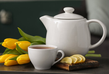 Cup of hot brewed tea, slices of lemon, pot and tulips on table against black blurred background