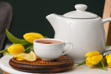 Board with cup of hot brewed tea, slices of lemon, tulips and pot on table against black blurred background