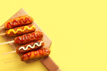Wooden board of tasty corn dogs with different sauces on yellow background