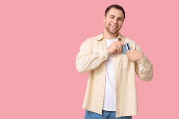 Young man putting credit card in pocket on pink background