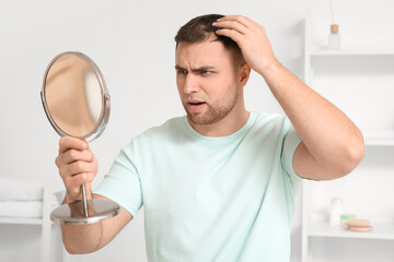 Worried young man with hair loss problem and mirror in bathroom