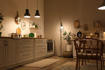 Interior of modern kitchen with glowing lamps, white counters, dishware and chair at evening