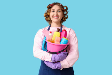 Young woman with hair curlers and cleaning supplies on blue background