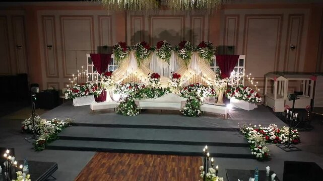 A cinematic drone footage capturing a grand Pakistani wedding stage with intricate decorations, floral arrangements, lush greenery, and soft back lights, featuring white sofa seating , drone camera vi