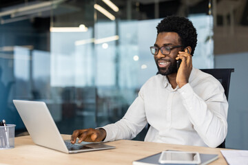 A cheerful African American man wearing glasses works on his laptop while talking on his cell phone...