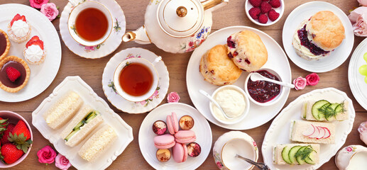 Afternoon tea table scene against a light brown wood banner background. Vintage tea set, finger sandwiches, scones, and desserts. Top view.
