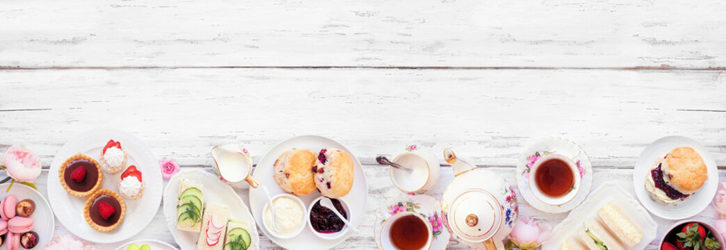 Afternoon tea bottom border against a white wood banner background. Vintage tea set, finger sandwiches, scones, and desserts. Top view with copy space.