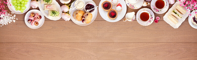 Afternoon tea top border against a light brown wood banner background. Vintage tea set, finger sandwiches, scones, and desserts. Above view with copy space.