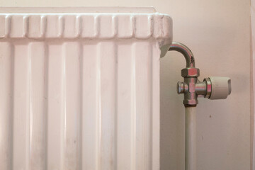 A close-up of a white cast iron radiator with a metal pipe and valve against a beige wall. The...